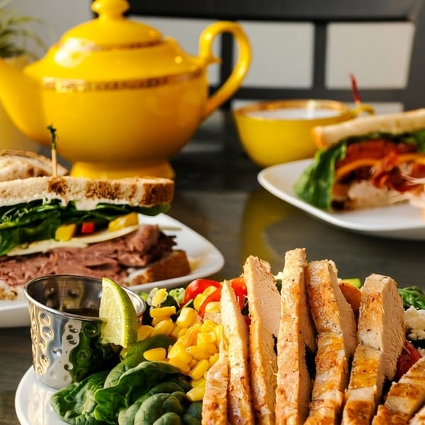 Plates of sandwiches, a chicken salad, and a yellow teapot with teacups on a table.