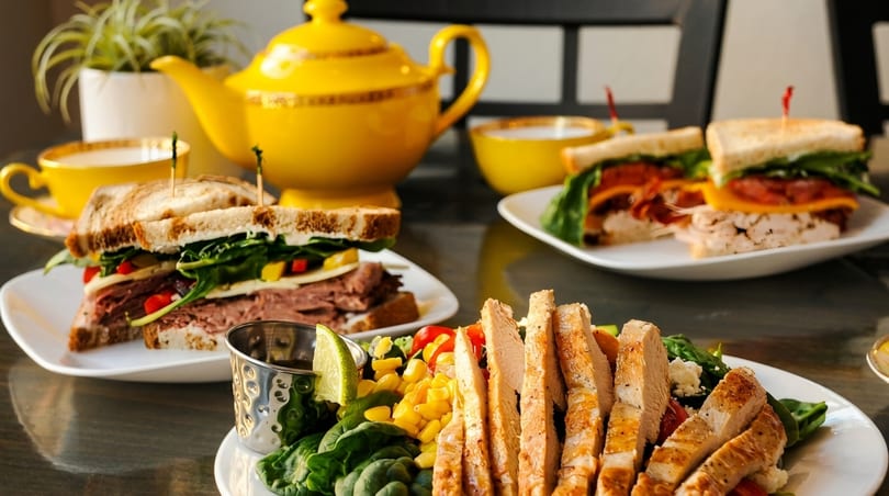 Plates of sandwiches, a chicken salad, and a yellow teapot with teacups on a table.