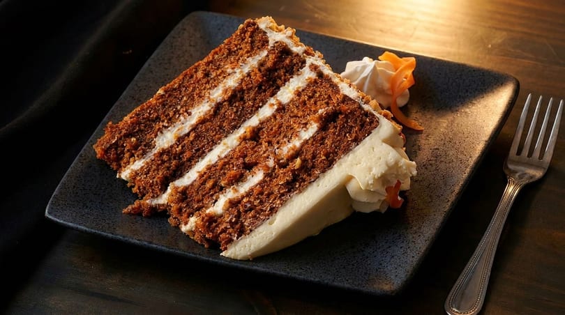 A slice of carrot cake with cream cheese frosting and a fork on a dark plate.