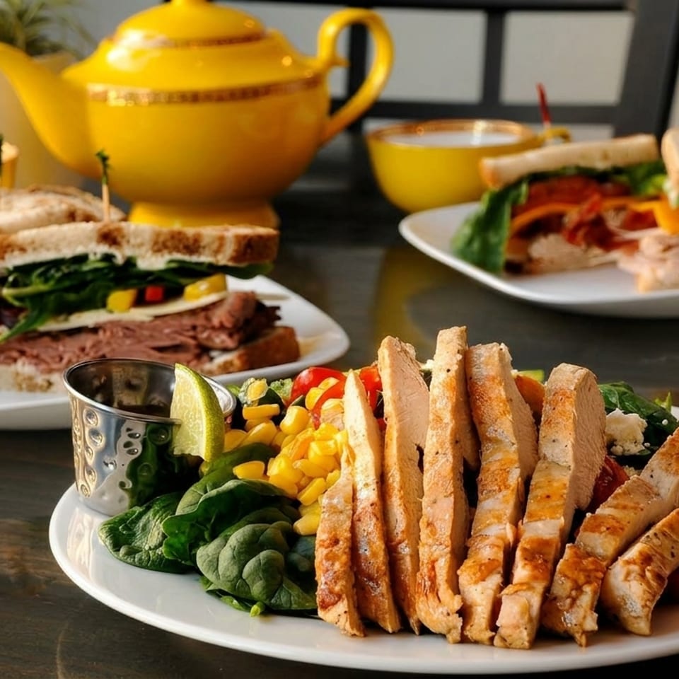 Plates of sandwiches and a grilled chicken salad with a yellow teapot and cups in the background.