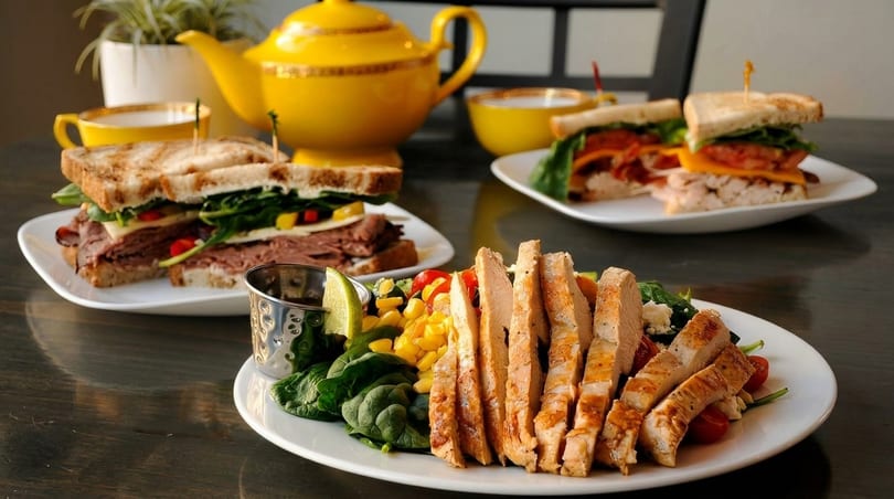 Plates of sandwiches and a grilled chicken salad with a yellow teapot and cups in the background.
