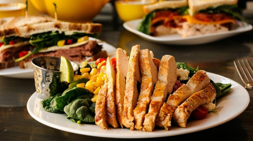 Sliced grilled chicken breast with spinach, corn, and cherry tomatoes, with sandwiches in the background.